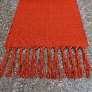 May include: Orange burlap table runner with a fringe border. The runner is made of a natural material and has a rustic look.