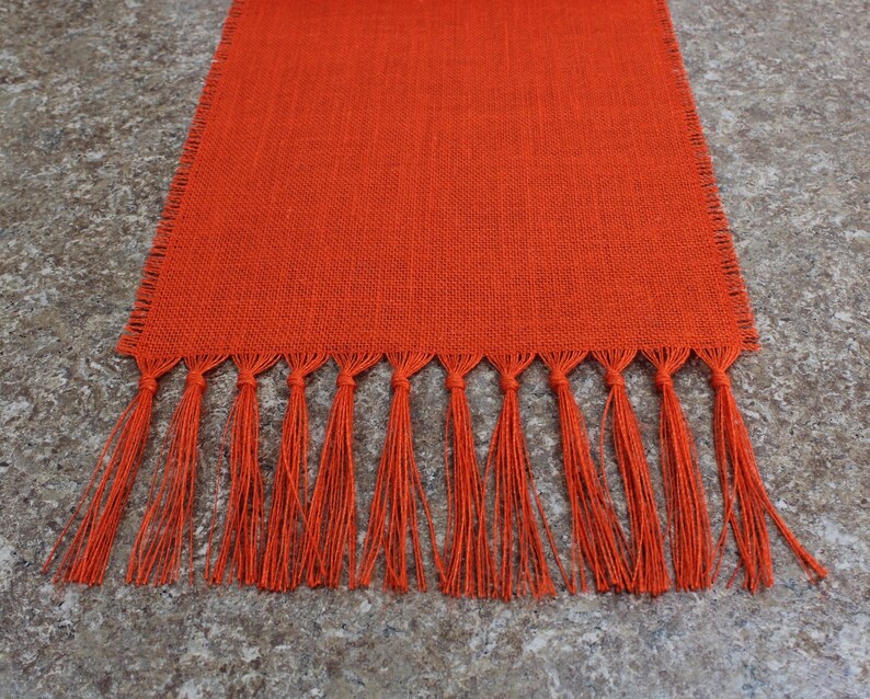 May include: Orange burlap table runner with a fringe border. The runner is made of a natural material and has a rustic look.