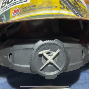 Może przedstawiać: Czarny kask z brązowym daszkiem i czarnym urządzeniem bezpieczeństwa z srebrnym logo z napisem "Roughneck". Kask ma naklejkę z napisem "Marathon Petroleum Company LP".