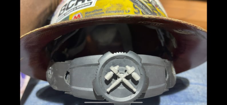 Może przedstawiać: Kask ochronny z brązowym sk&oacute;rzanym daszkiem i ż&oacute;łtym wierzchołkiem. Kask ma naklejkę z napisem "Marathon Petroleum Company LP". Kask jest noszony przez osobę i widoczny jest czarny pasek z szarym emblematem dw&oacute;ch skrzyżowanych kilof&oacute;w.