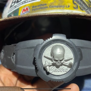 Puede incluir: Un casco de seguridad negro con un diseño de calavera y huesos cruzados en el lateral. La calavera es plateada y los huesos cruzados son negros. El texto "Danger" está escrito debajo de la calavera.