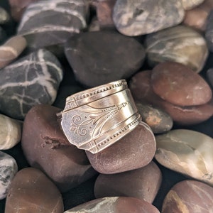 May include: A silver spoon ring with an ornate design, resting on a bed of smooth, gray and brown stones.