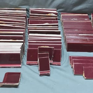 May include: A collection of burgundy-colored ceramic tiles, arranged in stacks and rows. The tiles vary in size and shape, including square, rectangular, and curved edge pieces. Some tiles have a glossy finish, while others appear matte.