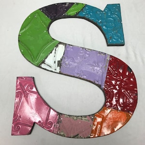 May include: A colorful metal letter S, made from repurposed tin tiles. The letter is painted in shades of green, purple, pink, red, orange, and blue. Each tile has a unique pattern and texture.