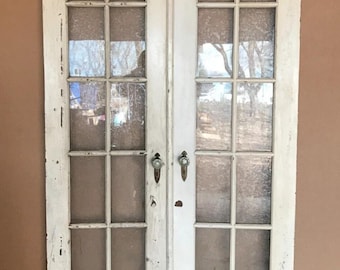 May include: A pair of antique white-painted doors with multiple glass panes. The doors have ornate handles and show signs of wear, with peeling paint and visible wood grain. The glass panes offer a glimpse of the outside.