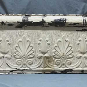 May include: An antique, rectangular architectural element with a distressed, off-white finish. The piece features a repeating pattern of stylized leaves and scrolls, with areas of exposed dark metal. The overall aesthetic suggests a vintage or reclaimed item.
