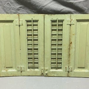 May include: Two-panel, light green wooden shutters with a distressed finish. Each panel features a rectangular design with a central section of vertical slats. Hinges and a latch are visible.