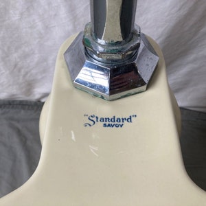 May include: Close-up of a white porcelain toilet tank with a chrome flush handle. The tank has a blue and white logo that reads "Standard" Savoy.