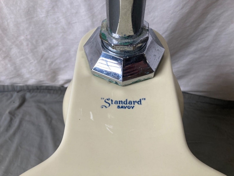 May include: Close-up of a white porcelain toilet tank with a chrome flush handle. The tank has a blue and white logo that reads "Standard" Savoy.