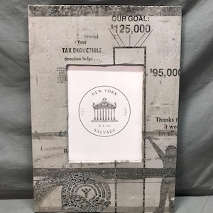 May include: Rectangular photo frame with a distressed newspaper print design. The frame has a white interior and features the text "NEW YORK 8 X 10 SALVAGE" and "OUR GOAL: $125,000".