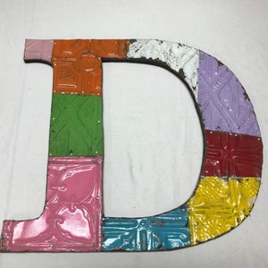 May include: A colorful metal letter D made of repurposed tin tiles. The letter is painted in shades of pink, orange, green, yellow, blue, and red.