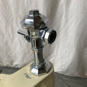May include: Close-up of a chrome toilet tank fill valve with a white porcelain toilet bowl. The toilet bowl has the text "Standard" and "Savoy" printed on it.
