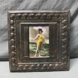 May include: Antique framed image of a woman in a yellow swimsuit standing on a beach. The frame is square, with a dark, textured finish and a beaded border. The image is a vintage photograph.