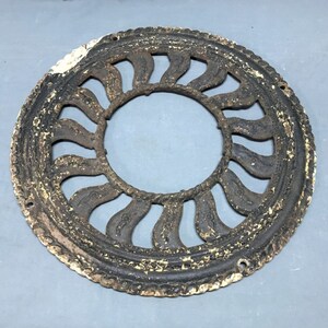 May include: A round, black cast iron vent cover with a decorative design of curved, pointed shapes. The cover has a distressed, aged look with white paint flaking off.