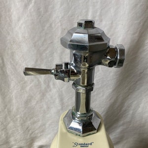 May include: Close-up of a chrome toilet tank fill valve with a lever handle. The valve is attached to a white porcelain toilet tank with the text "Standard" Savoy printed on it.