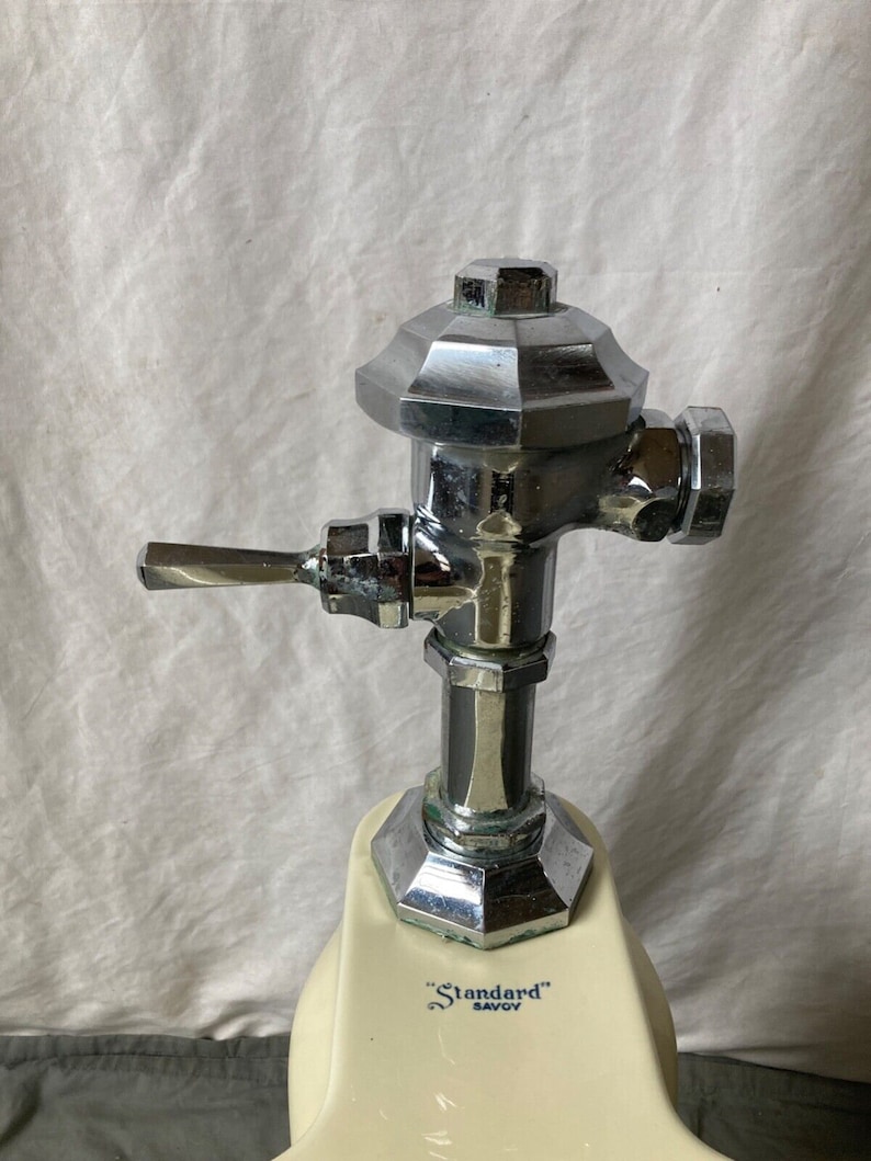 May include: Close-up of a chrome toilet tank fill valve with a lever handle. The valve is attached to a white porcelain toilet tank with the text "Standard" Savoy printed on it.