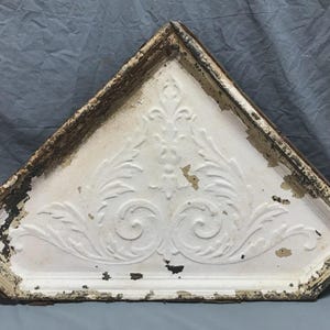 May include: A white, triangular architectural element with a decorative, embossed floral design. The edges are framed with a dark, weathered trim. The paint is chipped, giving it a vintage appearance.