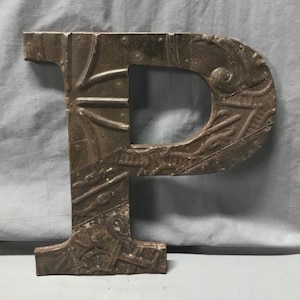 May include: A large, weathered metal letter "P" with a distressed, industrial aesthetic. The letter is a dark, muted brown with riveted details and embossed patterns. The design has a vintage, rustic feel, suitable for wall decor.