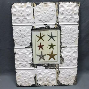 May include: White framed picture with a distressed, aged look. The frame is made of tiles with a raised floral design. The picture inside is of a vintage illustration of starfish.