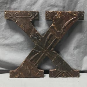 May include: A large, decorative metal letter "X" in a rustic, distressed finish. The letter is brown with darker accents and embossed floral and geometric patterns. The metal has a weathered appearance.