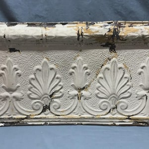 May include: An antique, off-white decorative architectural molding with a repeating leaf and scroll design. The molding has a textured surface and shows signs of age with distressed paint and dark edges. A vintage home decor item.
