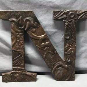 May include: A large, decorative metal letter "N" with a distressed, antique finish. The letter is brown with embossed floral and geometric patterns, and riveted metal accents. The overall style is rustic and vintage.