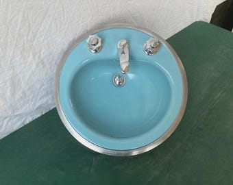 May include: A vintage-style, oval-shaped bathroom sink in a light blue color. The sink has a chrome faucet and two chrome knobs. The rim of the sink is silver. The sink is a classic design for a bathroom.