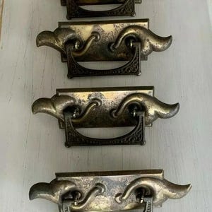 May include: Four antique brass drawer pulls with ornate designs. Each pull has a rectangular base with a decorative handle and a curved, wing-like design on each side.