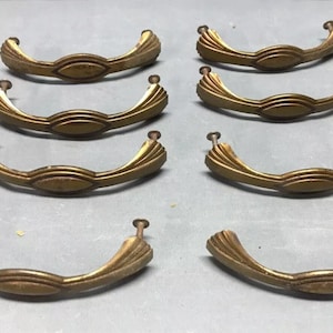 May include: Eight gold-toned metal cabinet door pulls with a decorative, curved design. The pulls have a slightly aged finish.