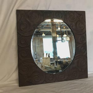 May include: Square brown mirror with an oval mirror in the center. The frame has a textured, swirling design. The mirror reflects a room with windows and hanging lights. A decorative wall mirror.