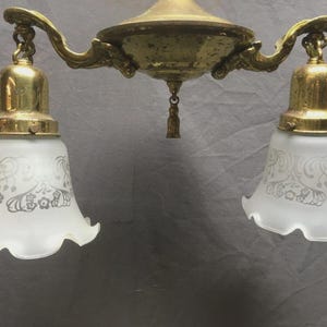 May include: Antique brass chandelier with two frosted glass shades. The shades have a floral design. The fixture has a decorative center and curved arms. The light fixture is a vintage style.