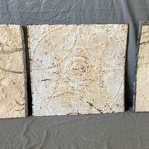 May include: Three antique, square, white tin ceiling tiles with embossed floral and geometric patterns. Each tile has a central circular design and is weathered with age, showing signs of rust and wear. The tiles are arranged side by side.