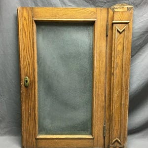 May include: A vintage wooden cabinet door with a frosted glass panel. The door has a keyhole and is made of light brown wood.