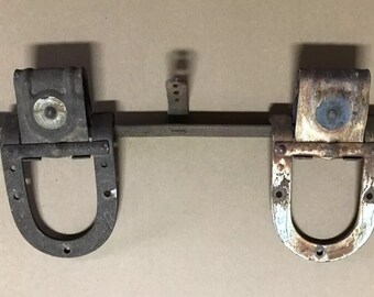 May include: Two antique metal door hinges with a horseshoe shape and a round metal plate in the center. The hinges are attached to a metal bar.