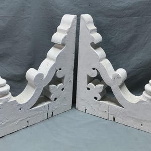 May include: Two white, ornate wooden architectural brackets. Each bracket features a decorative scroll design, a floral motif, and a tiered finial. The brackets are painted white and appear to be aged.