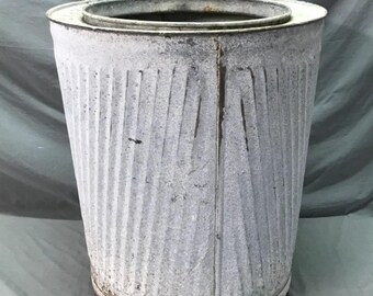 May include: A vintage, galvanized metal trash can with a white, weathered finish. The can has vertical ribbed detailing and a circular rim. The bottom rim shows signs of rust, adding to its antique appearance. The can is cylindrical and tapers slightly towards the base.