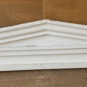 May include: A white, wooden architectural pediment with a triangular top and rectangular base. The pediment has a layered design with decorative molding and appears to be aged or distressed. The piece is painted white and rests on a wooden surface.