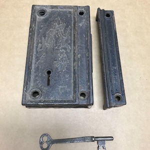 May include: Antique black cast iron door lock with a key. The lock has a keyhole and two sets of screws. The key is also black metal.