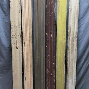 May include: Six weathered wooden architectural trim pieces in varying colors, including cream, tan, olive green, burgundy, yellow, and off-white. The pieces have vertical grooves and peeling paint, suggesting an antique or reclaimed aesthetic.