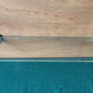 May include: A clear glass shelf with metal supports. The shelf is rectangular with a beveled edge. The metal supports are silver and have a decorative starburst design. The shelf is mounted on a wooden surface.