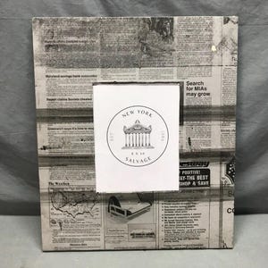 May include: A rectangular photo frame with a newspaper print design. The frame has a white insert with the text "NEW YORK 8 X 10 SALVAGE" and a graphic of a building. The frame is designed to hold an 8x10 inch photo.