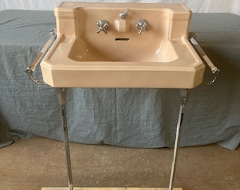 May include: A vintage peach-colored porcelain bathroom sink with chrome fixtures and a chrome towel rack. The sink is mounted on a chrome stand with a wooden base.