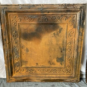 May include: A vintage, rusty metal ceiling tile with an ornate, decorative design. The tile is a square shape with a raised border and a central design of swirling scrolls. The tile is a dark brown color with a patina of rust.