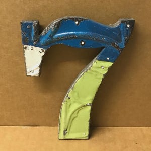 May include: A vintage metal number seven sign. The number is painted in blue, white, and green with a distressed finish. The sign is attached with small rivets and has a rustic, industrial aesthetic.