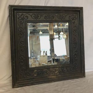 May include: Square, dark-toned mirror with an ornate, scrollwork-detailed frame. The mirror reflects a room with industrial elements, including windows and hanging lights. The frame has a slightly distressed appearance.