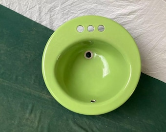 May include: A bright green ceramic bathroom sink with three holes for faucets. The sink is round and has a smooth, glossy finish.