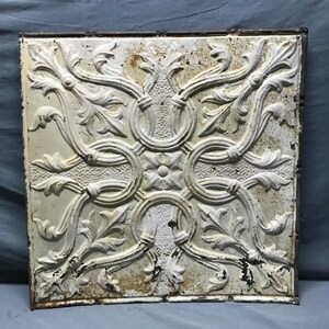 May include: Square, off-white metal ceiling tile with a detailed, raised floral and scrollwork design. The tile has a distressed, aged appearance with rust-colored spots and a dark brown metal frame. The design features a central flower and repeating circular patterns.