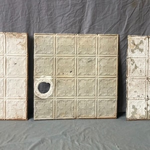 May include: Three white tin ceiling tiles with a raised square pattern. The tiles are slightly different shades of white and have some wear and tear. One tile has a hole in the center.