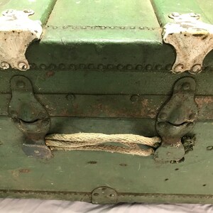 May include: A vintage green metal trunk with rope handles and white metal clasps. The trunk has a worn, distressed look.