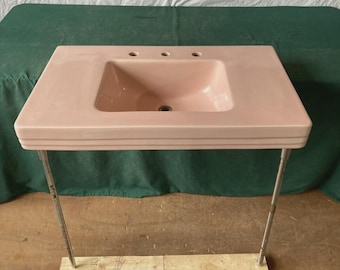 May include: A rectangular pastel pink bathroom sink with a central basin and three faucet holes along the back edge. The sink features a tiered, stepped front design and is supported by two thin, vertical metal legs attached to a wooden base.